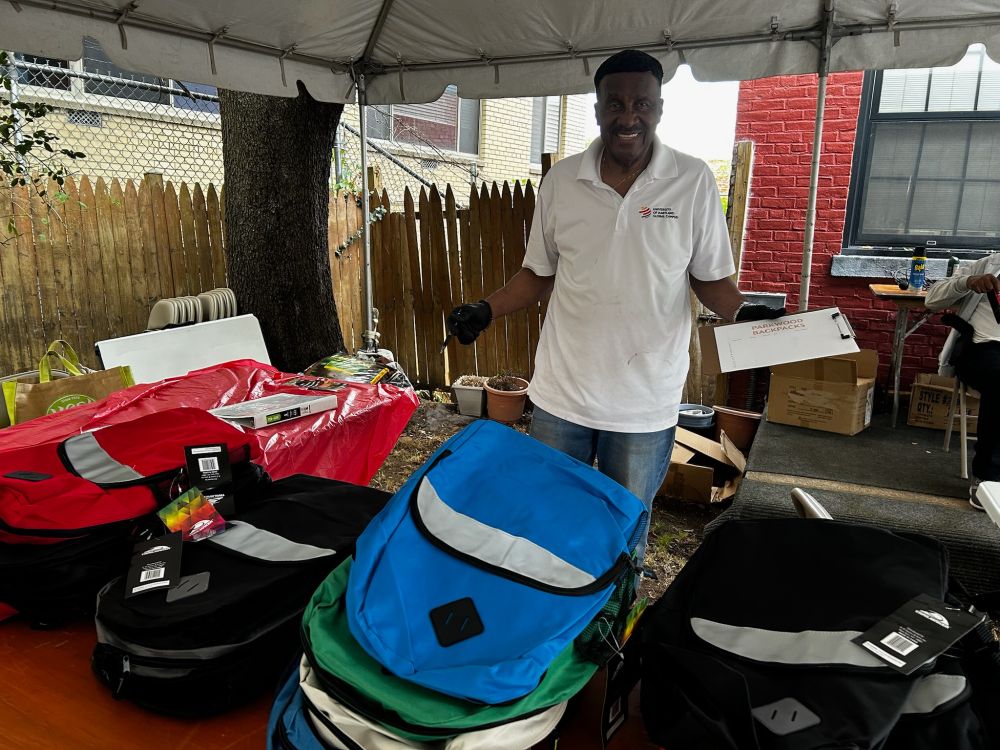 A man stands in back of a table full of backpacks under a tent at the Parkwood Place block party