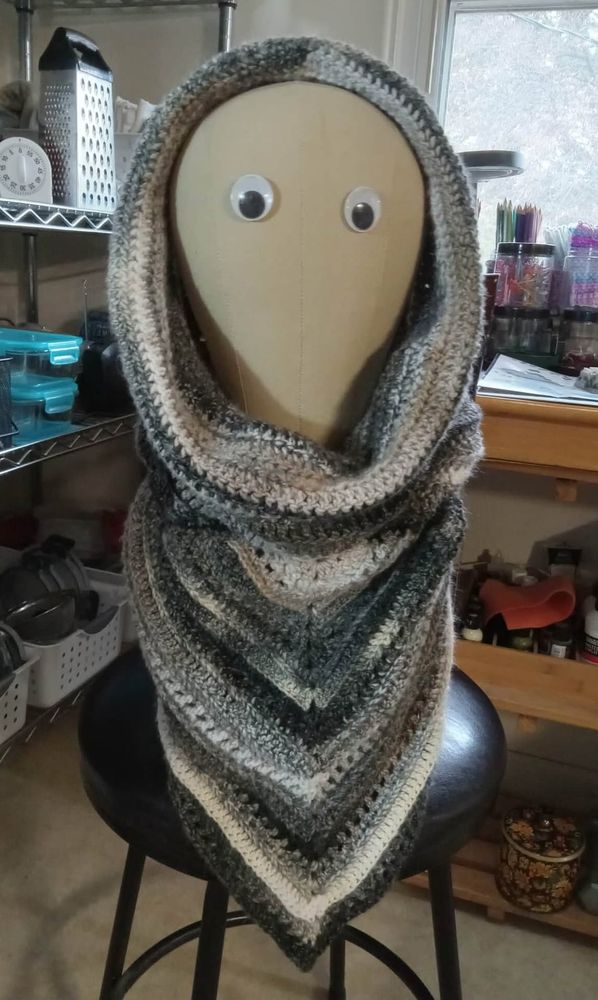 Image of a crocheted, hooded scarf modeled on a googly-eyed mannikin head, and displayed with my junky kitchen corner in the background. The scarf is in shades of gray, beige, and white.