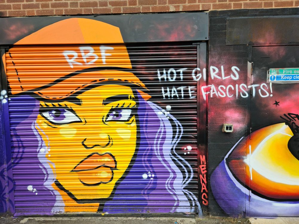 A shop shutter painted in street art style with a bright yellow person with long purple hair, purple eyes and an orange cap with RBF on in white. The tag reads Hot girls hate fascists. 