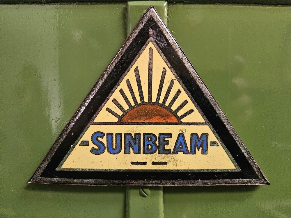 A pale yellow triangular metal sign which reads "Sunbeam" in blue letters with a sun rise graphic above it. It's on green metal panels.