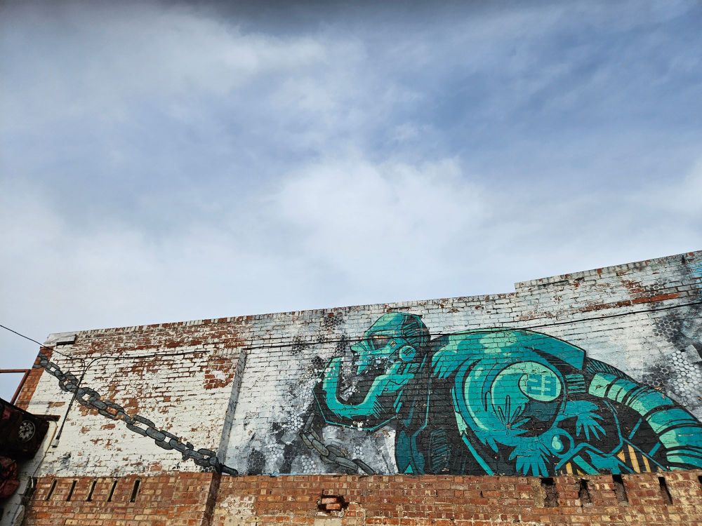 The bottom half of the picture shows a brick wall painted with a large hulking turquoise robot in profile and a chain from the robot to the edge of the wall. The robot is looking up at the blue sky above, it has a large open jaw, and an angry expression.  