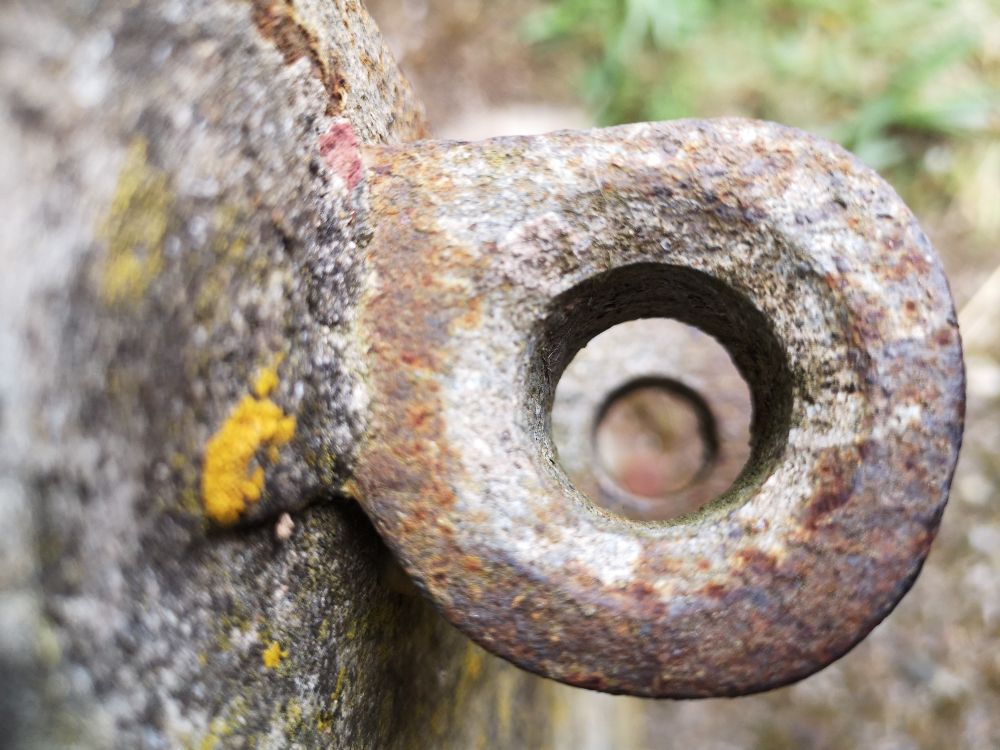 Close up of an empty round rusty iron hinge on a stone mount. Through the hole in the centre you can see another hinge and circular hole. There is yellow lichen on the stone wall, and green plants in the background. 