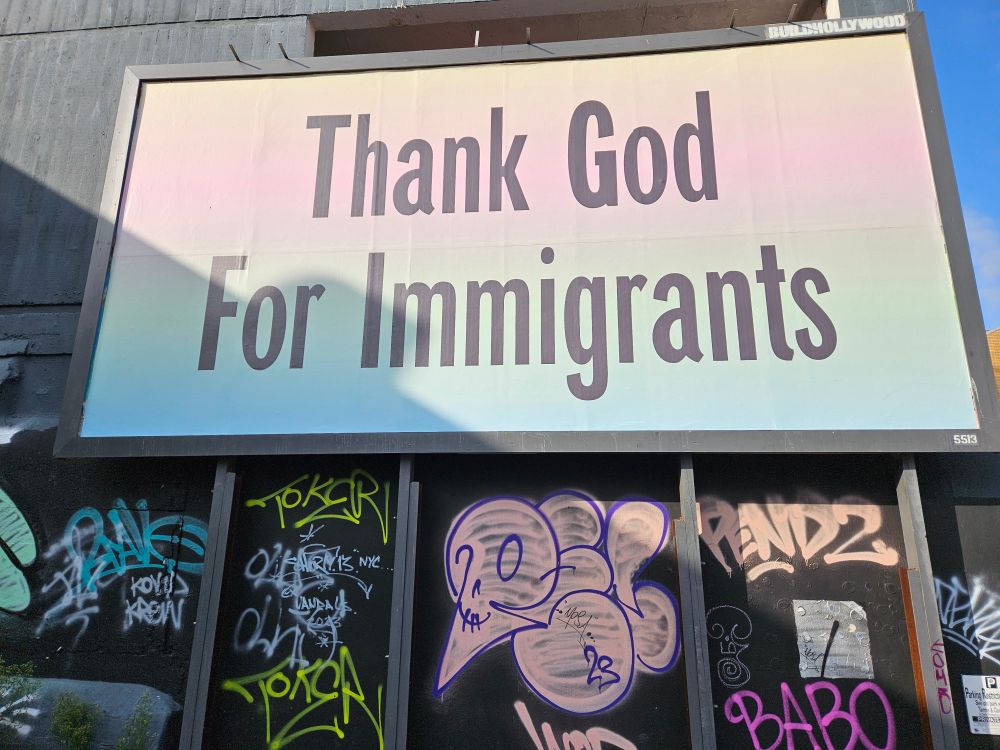 A billboard in pink and turquoise with "Thank God for Immigrants" written in black, there are graffitti tags under the billboard. 