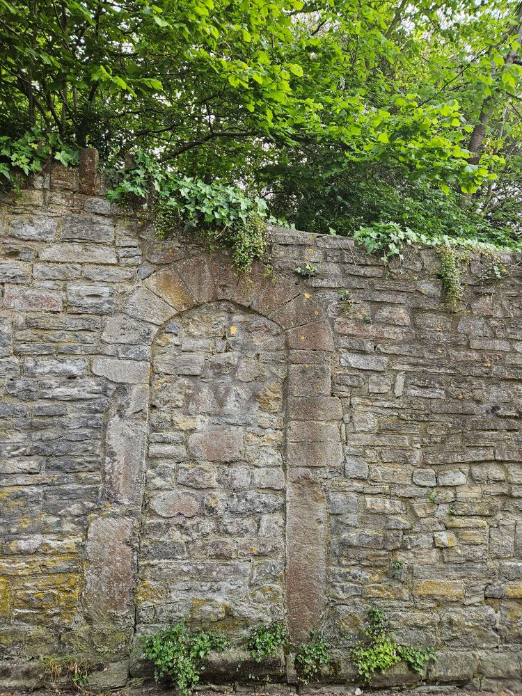 An old great stone wall, with the outline of a gateway filled in with similar stone. There's ivy growing on top and trees visible above.