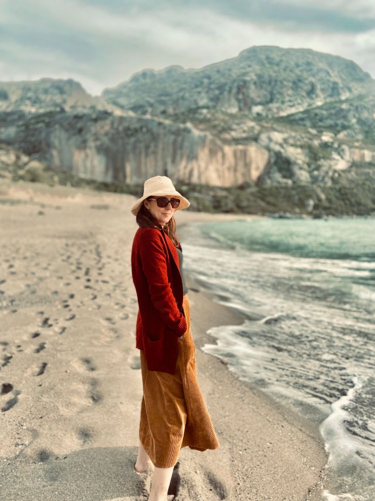 Woman (me!) standing on a beach in Crete. You can see waves coming in to the right and dramatic cliffs in the background. There are footprints in the sand. The woman is barefoot, wearing a long dark red cardigan, a long tan corduroy skirt, sunglasses and a dorky straw hat. The photographer (aged 13) has given the photo  a vintage-looking tint.