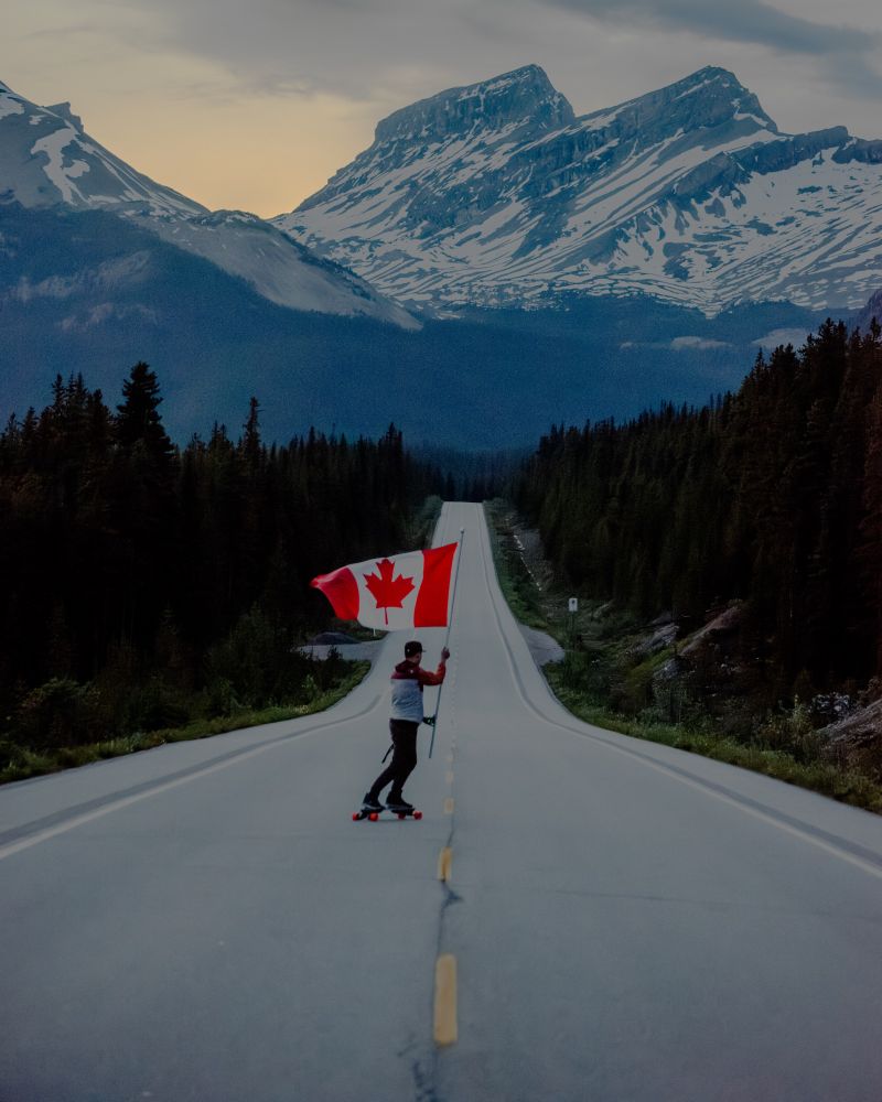 A man skates down a stretch of road in the Rocky Mountains waving a Canadian flag 