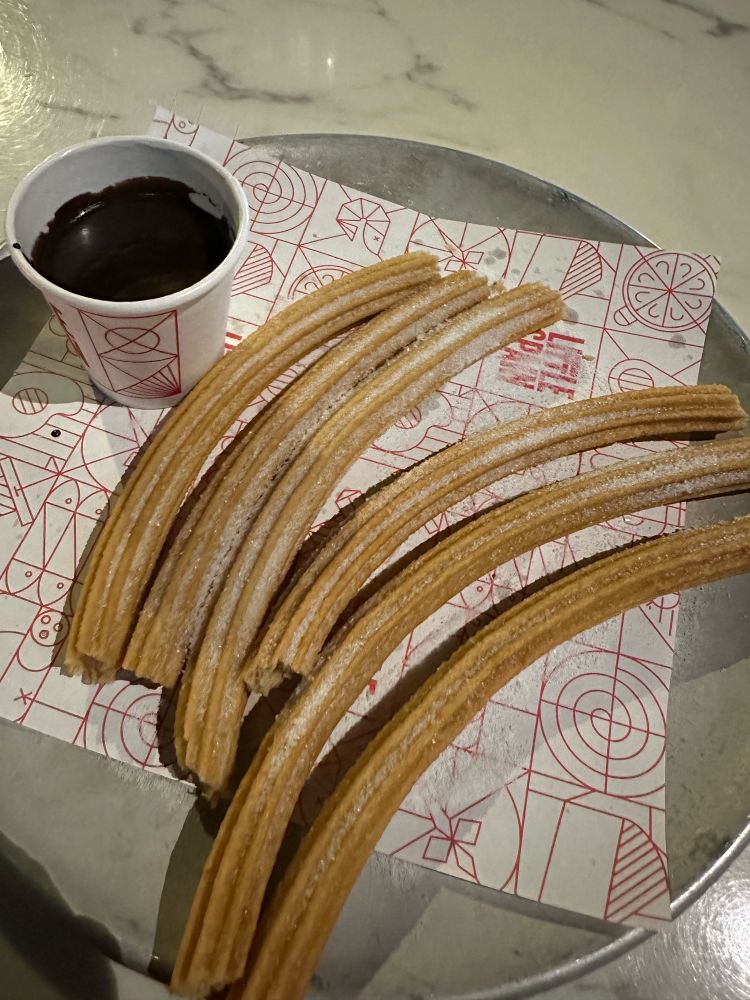 A round tray with a paper on it and six churros on top, one missing a bit at the bottom. A small cup of thick, hot chocolate sits beside the churros, waiting for them to be dipped into it. All of this is on an outdoor standing table.