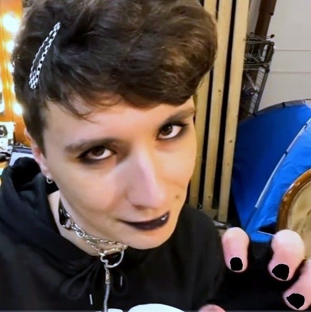 A selfie taken by Dan Howell, a white man with curly brown hair and brown eyes. He is wearing a black hoodie, a chain choker, and black fingerless gloves. He is holding a hand up like a cat paw. He has a black and white checkered barrette in his hair. Eyeliner, black eyeshadow, black lipstick, and black nail polish have been edited on.