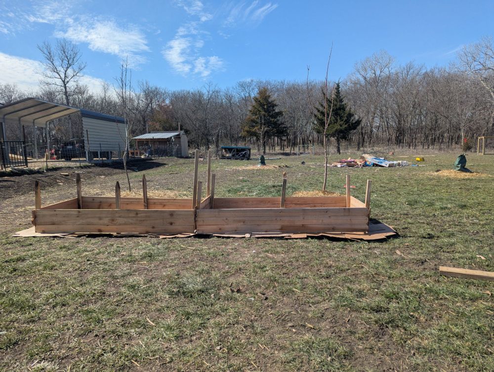 Two raised garden beds, built from cedar fence slats, with 12 one inch posts sticking up for support. It's a little wonky, but it will work to grow things in it. In the background, there's a beautiful blue sky with thin white clouds, and a forest of leafless trees.
