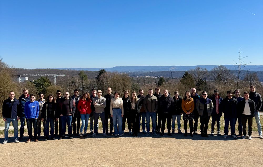 A wide shot of approximately 30 individuals standing in a line, posing for a group photograph outdoors. The background shows a clear blue sky, trees, and a distant cityscape or hills.