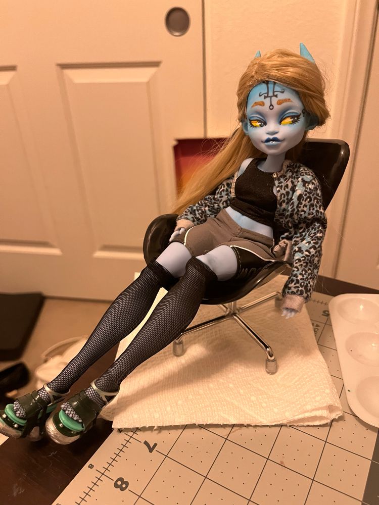 a photo of an 11-inch fashion doll sitting on a doll-sized chair on a human-sized workspace. she is a blue-skinned, horned devil, with blonde hair, yellow eyes, and a smug look on her face. she’s wearing casual streetwear that shows off plenty of plastic skin.