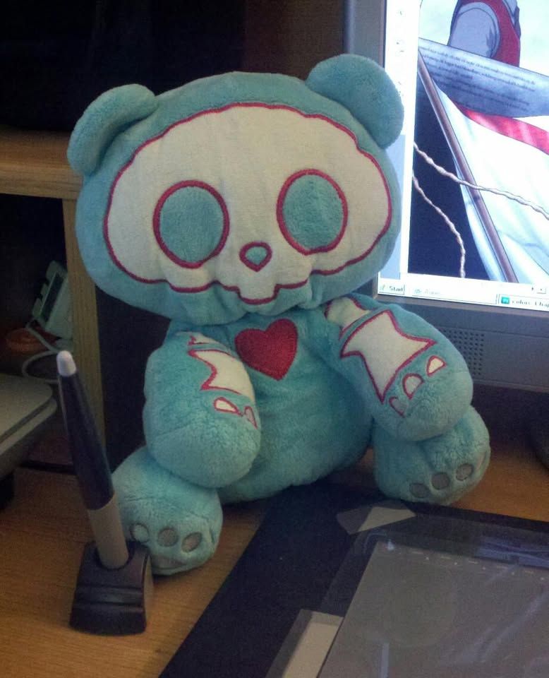 Photo of my old desk from 2011, back when I started drawing my webcomic, MONSTER SOUP. On the desk is a plush light blue teddy bear with a skeleton pattern, and a red heart on the chest. An Intuos 2 drawing tablet is also seen.
