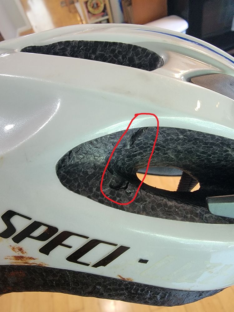 A closeup photo I took of my old bicycle helmet, focusing on the right side about halfway between the front and back, and about halfway from the bottom-most edge to the top peak.  Circled in red is a notched indentation in the black foam that forms the protective layer.  The indentation is about a quarter inch thick and runs mostly vertical for several inches (across a gap in the foam meant for ventilation), with the depth varying along the curved surface but reaching between a quarter and a half inch deep in places.