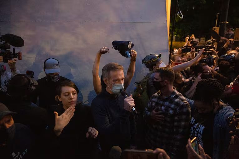 A protester holds a police hat over Ted Wheeler's head at a protest in 2020.