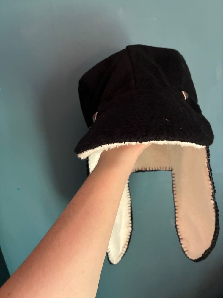 Front view of hat being held up by me. It is black with a white lining and two decorative studs on the front. It has a brim and the back goes down and has two flaps to cover my ears.