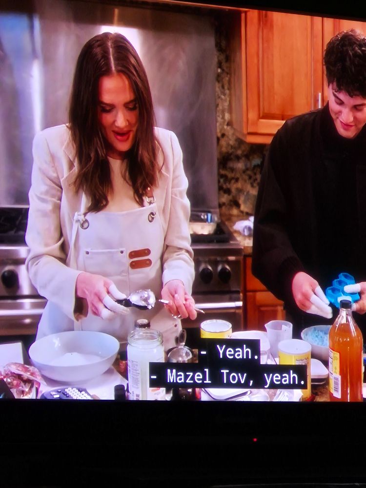 Meredith and Brooks Marks in the kitchen measuring ingredients for bath bombs with fiber condoms on most of their fingers. Meredith one has them on one hand but both of Brooks's hands have all 5 fingers covered. 