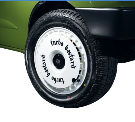 One of the front wheels of a Daihatsu car/minivan thing. It has a flat white hubcap on which are written, in a Gothic font, the words "turbo bastard".