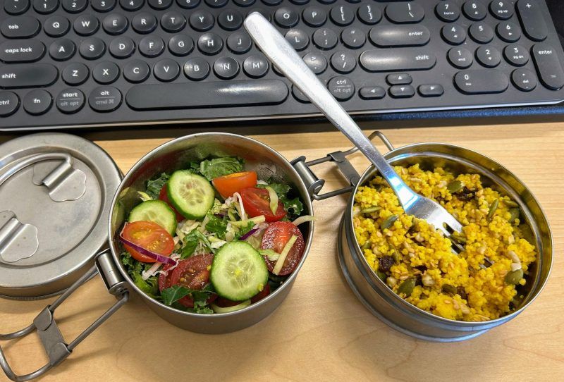 Two steel containers and a lid next to a keyboard on a desk. One container of cous cous and another of salad.