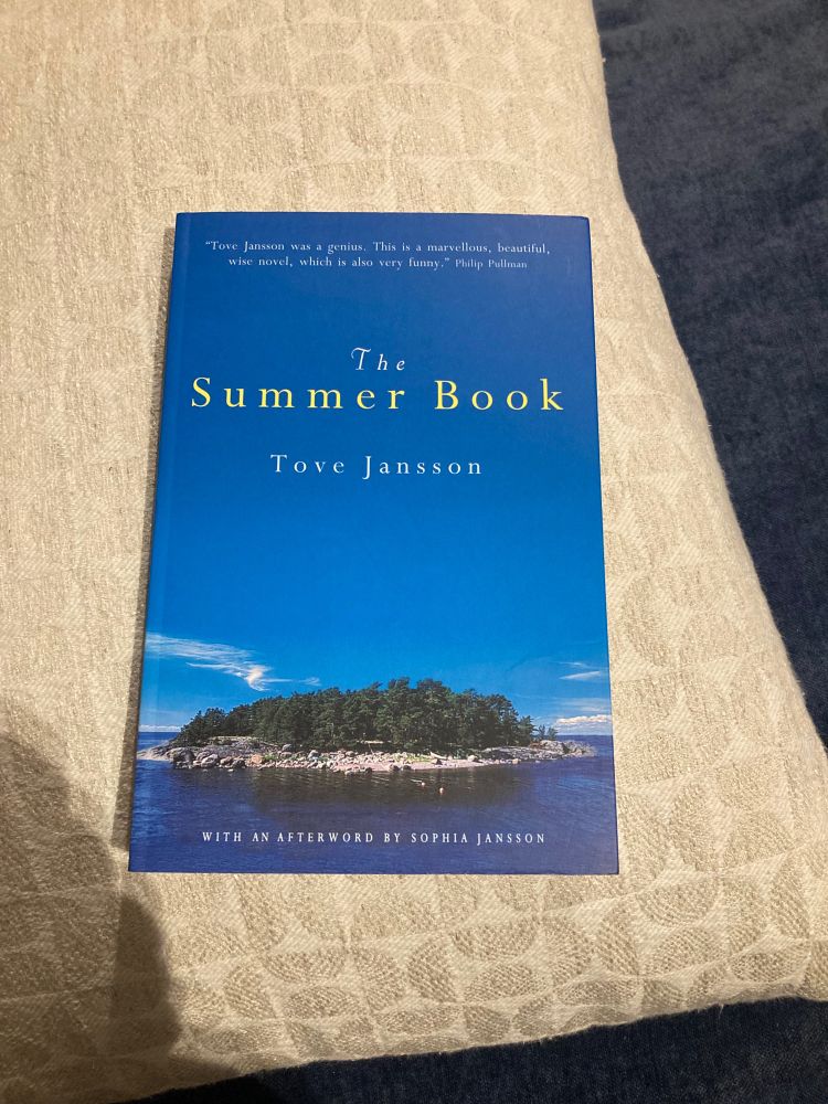 The Summer Book, by Tove Jansson. Cover shows a small, wild island, home to rocks and wind-bent trees. Cerulean sky above. Wispy summertime clouds above the horizon. The tiny Finnish island is surrounded by a cobalt blue sea.