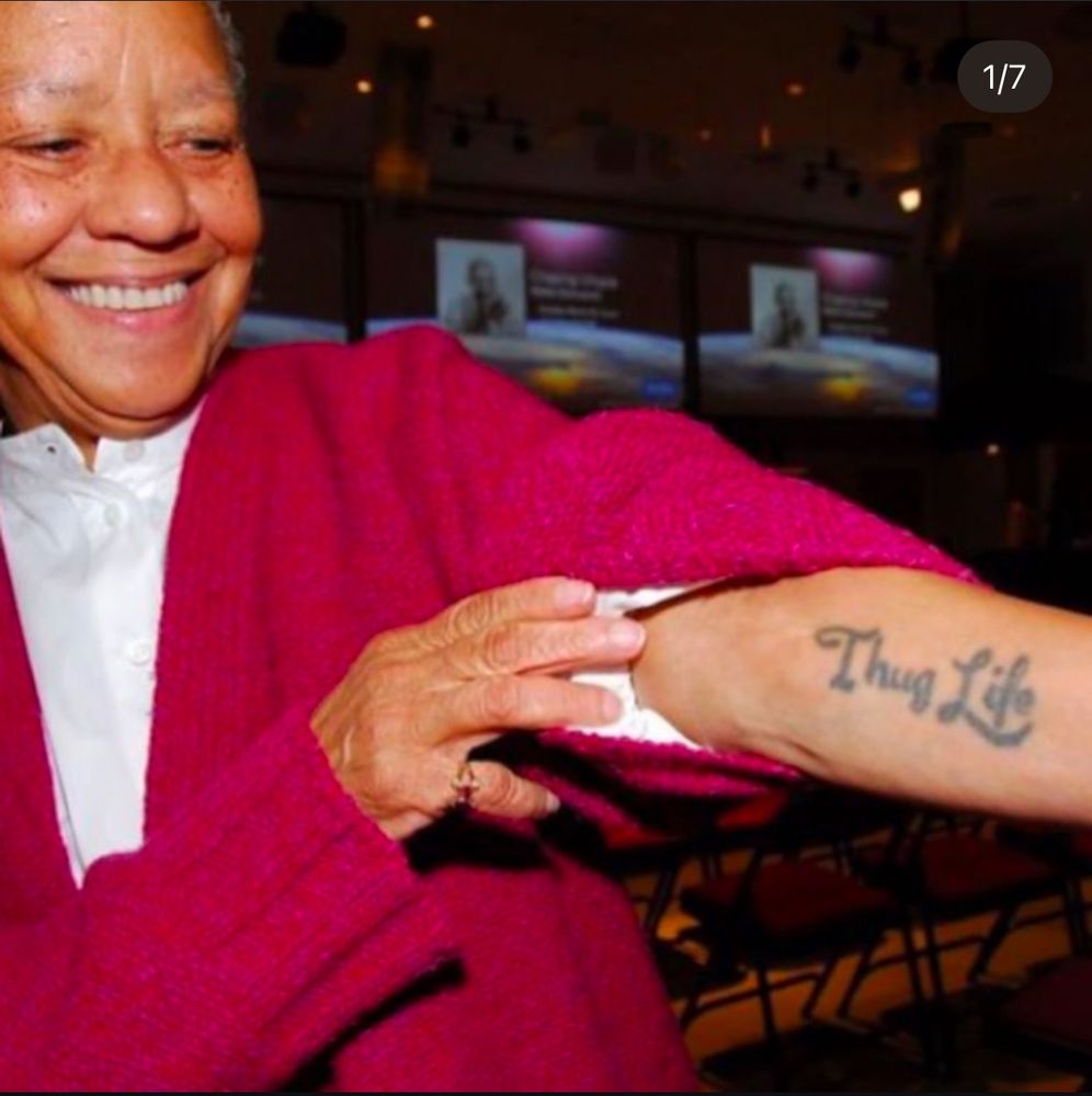 An older Nikki Giovanni smiling widely and showing a tattoo on her forearm that says, “Thug life” in cursive letters