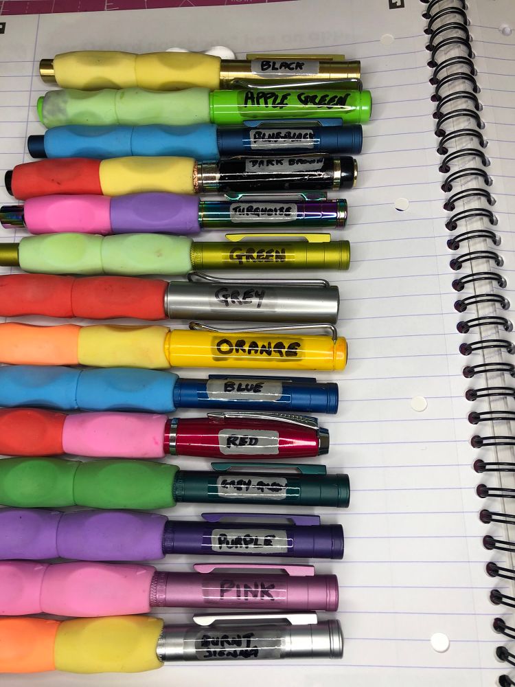 Fourteen fountain pens with two rubber triangle grips on each, in several different colours, sit in a row on a lined piece of A4 paper. Each of the pens has the colour of the ink that it carries written in permanent marker on the lid, varnished over with Army Painter Dark Tone.