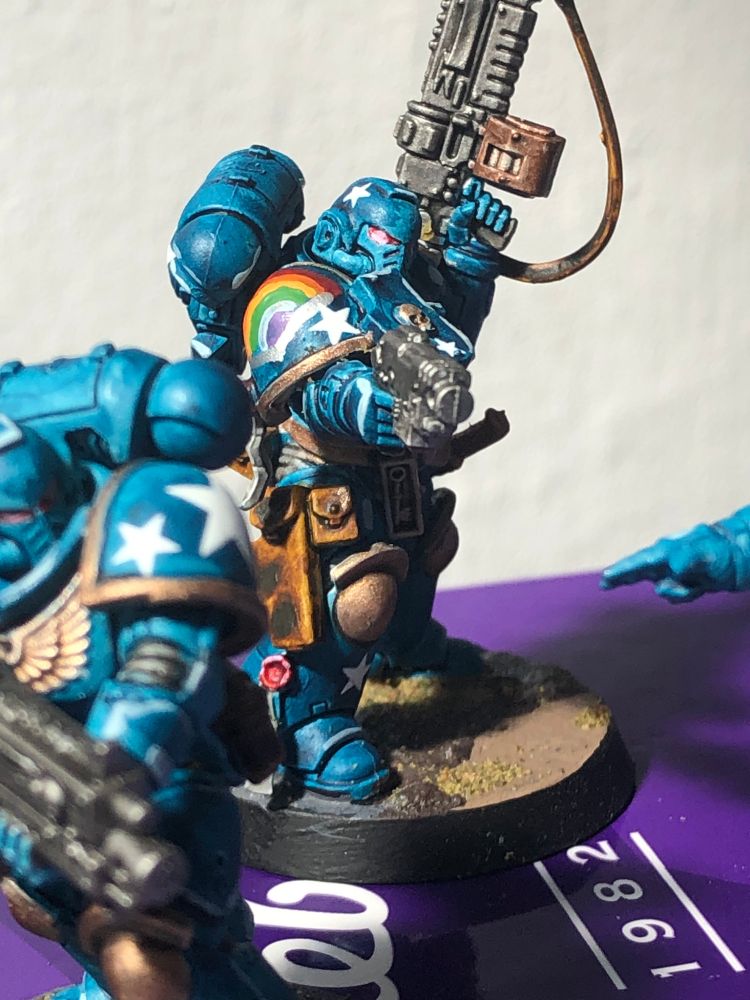 Pictured is a Space Marine Primaris Intercessor. It is painted in a shade of blue somewhere around PSG Blue with a Uranian Blue highlight (see: https://cssgradient.io/shades-of-blue/), with brass-gold accents and trim. Each model bears a stylised rainbow and the Chapter Symbol is an arrangement of white five pointed stars that differs from model to model.