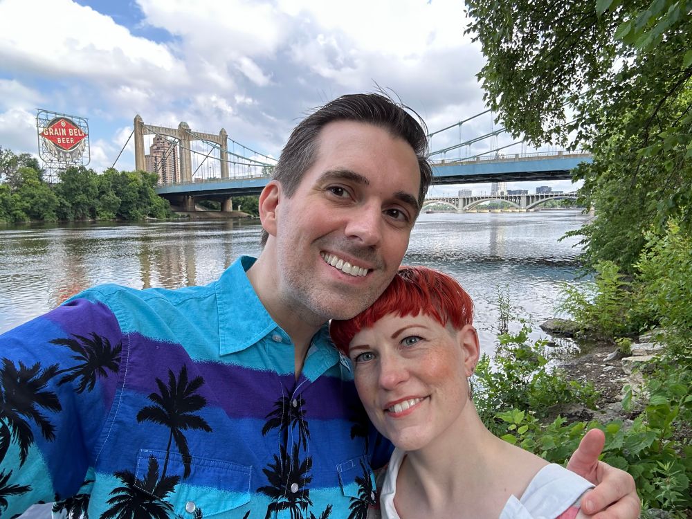 A white man wearing a tropical print shirt and a white woman posing in front of the river