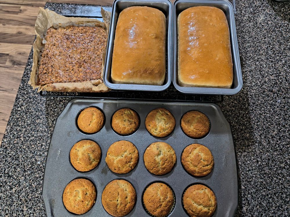 A pan of banana oat bars, two loaves of fresh baked bread, and a dozen banana muffins 