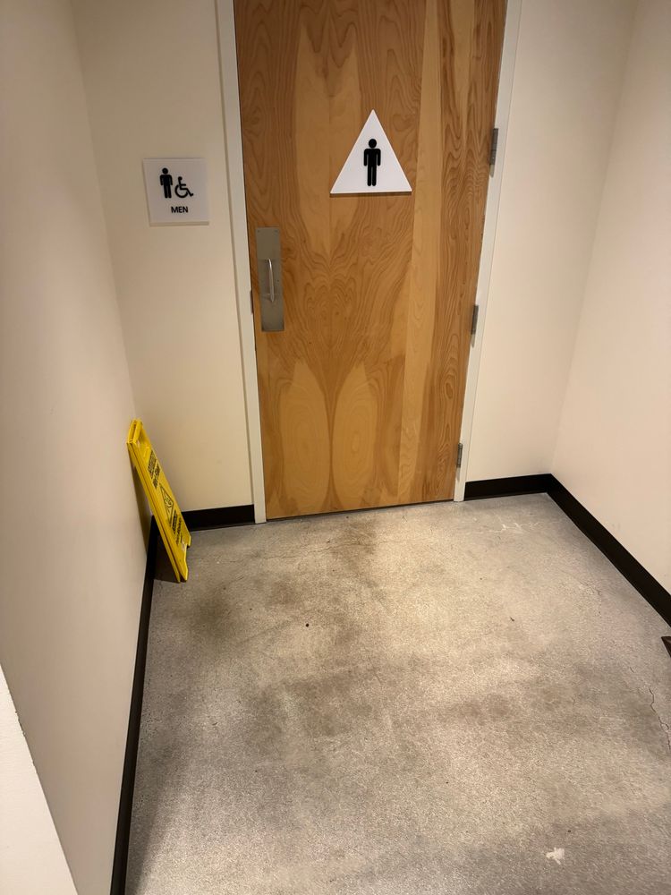 The floor in front of a men’s room door. The floor is darker in the spot where people stand to open the door. 