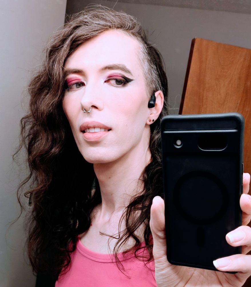 it's me! in a mirror selfie (so technically all the sides referenced here are wrong b/c theyre reversed in the reflection)! i'm a pale trans woman with wavy brown hair swept to the left side of my face and a side shave on the right. have a silver septum ring in my nose & two small studs, one red & one black, in my right earlobe. wearing a pink tank top. eyeshadow appears kind of a smeared pink, but it's *actually* a glittery maroon with a metallic glitter catching the light in such a way that it seems to match my top. also have nice & thick black eyeliner wings. lip color has worn off but thankfully my lips look great regardless. 