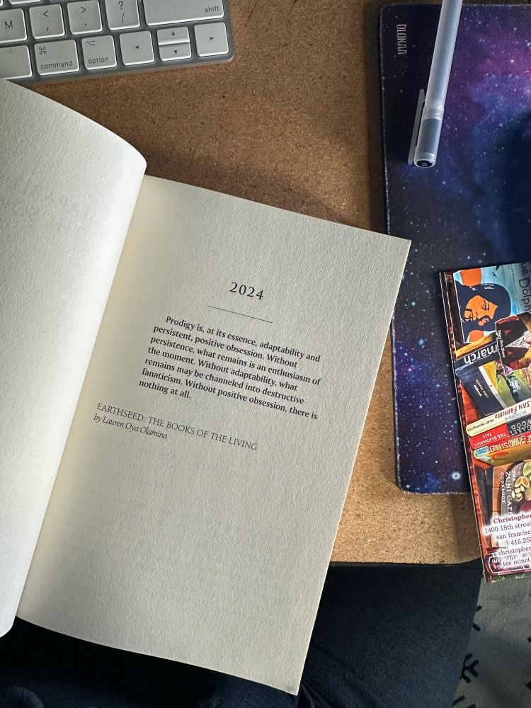 The opening page of Octavia Butler’s Parable of the Sower, which was published in 1993 and takes place in a speculative 2024. The book sits on a cork mat and there is a space printed mousepad and a bookmark from Christopher’s Books to the side. 