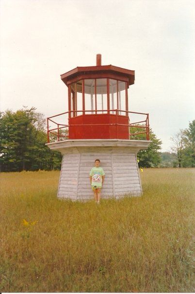 The top of a red and white lighthouse sits in a field while a young Scott wearing neon green stands against it.