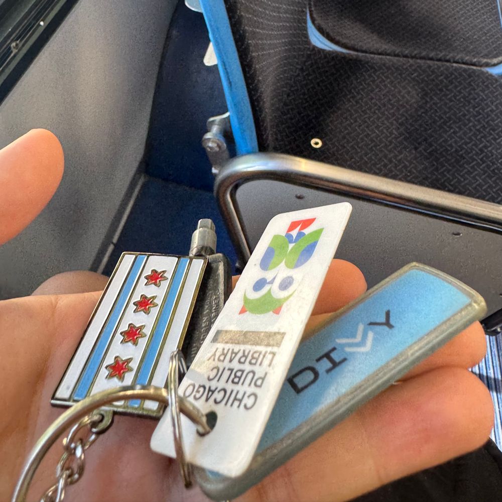 a photo of the additions to a big key ring all sitting in the end of my palm. the photo shows the hand in front of a bus chair. the key ring additions include an enamel keychain of the chicago flag, a hex key to a refrigerator, the keychain chicago public library library card, and the divvy key. 