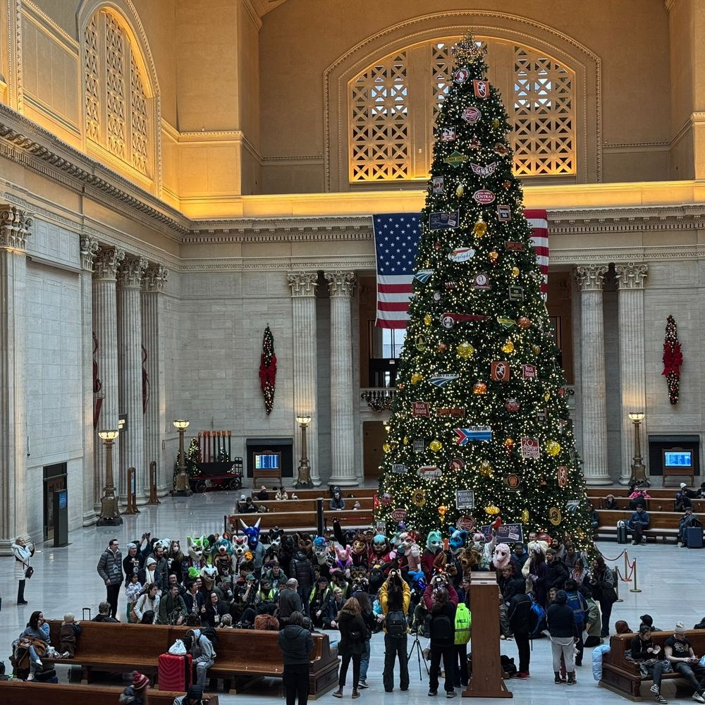A second photo of the Transit Oriented Animals people posing in front of a christmas tree in the Great Hall of Chicago Union Station, surrounded by benches of travelers. This photo is zoomed out and includes the entire christmas tree.