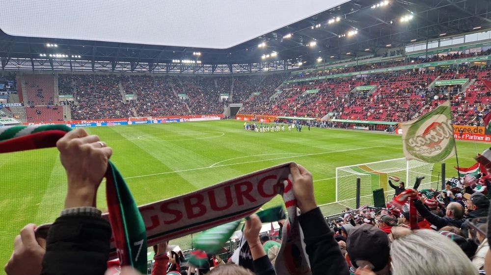 Aufnahme aus dem Stehblock des Augsburger Fußballstadions. Man sieht Fahnen, Menschen die Schals hochhalten und die einlaufenden Mannschaften. 