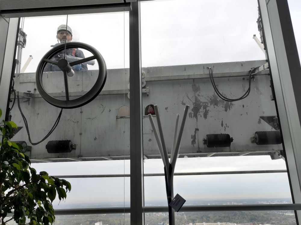 Foto eines Mannes mit Schutzhelm vor einem großen Fenster in großer Höhe. Er steht in einer Metallplattform mit hohem Rand.