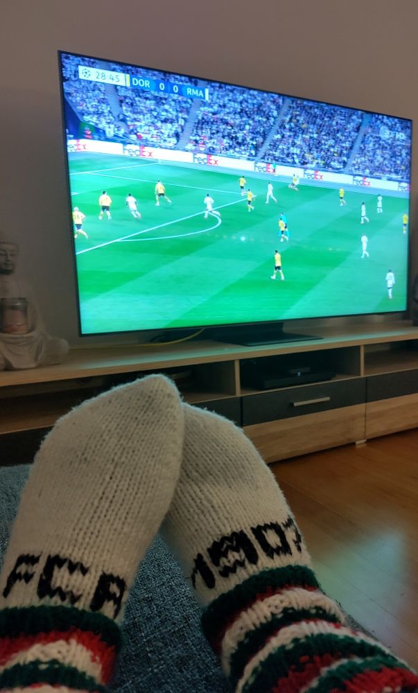 Vor einem Fernseher, auf dem das Champions League Finale läuft, sind zwei Füße in selbst gestrickten Socken zu sehen. Auf der weißen Wolle ist auf einem Fuß "FCA" aufgestickt, auf dem anderen "1907".
