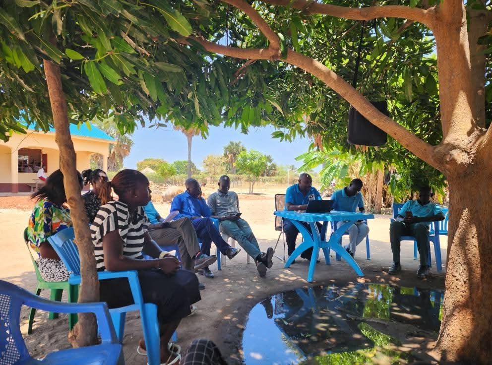 Teachers sit under a mango tree listening to their principal. 