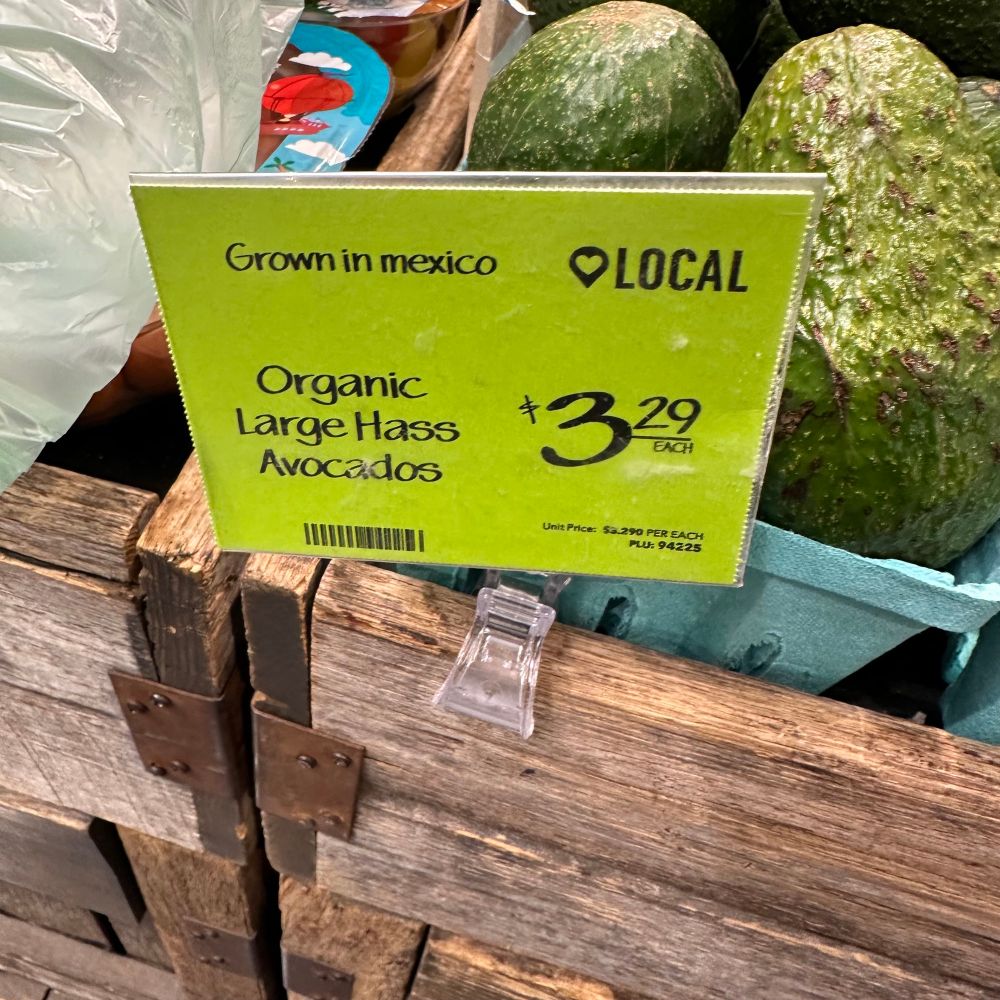 Sign at a store next to avocados saying “Grown in mexico” and “local” and shows the Organic Large Hass Avocados are $3.29 each