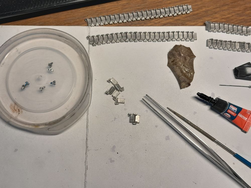 Four lengths of track for a 1:35 scale M3 Stuart tank, fitted together from metal link and pivot parts. A few separate link and pivot parts, ready for assembly. Modelmaker's tweezers and cylindrical reamer/file. A tube of superglue, a pin for wiping away excess glue, and a honeysuckle leaf to catch glue spills from the tube. The entire picture suggests that a time-consuming and fiddly task is in progress.