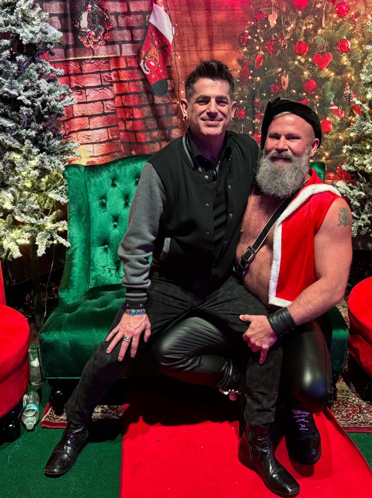 Man sitting on another man’s lap dressed as sexy Santa - shirtless in a red vest with white trim. 