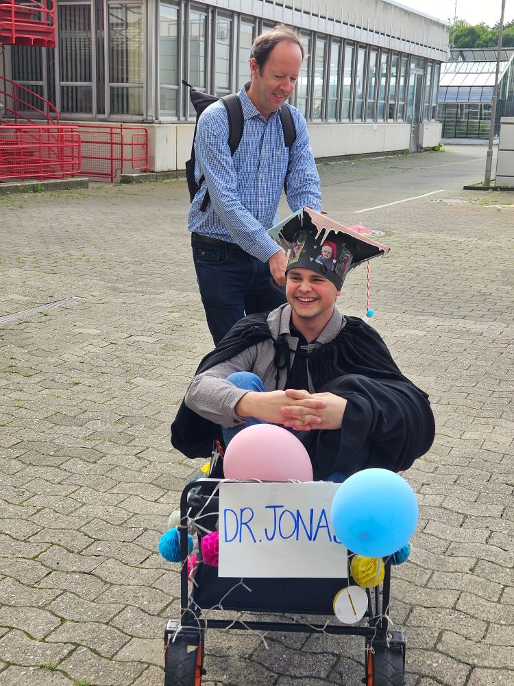Jonas Tebbe sitting in a small cart decorated with balloons and a sign that says "Dr Jonas". Jonas is wearing his graduation cap and a black gown. The cart is being pulled by his former supervisor Joe Hoffman. They are outside, some parts of Bielefeld University can be seen in the background