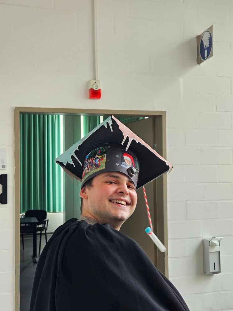 Jonas wearing a black gown and his graduation cap which is decorated with pictures and has a lab tube for a tassel. He is standing inside Bielefeld University in front of the room where he defended his PhD thesis
