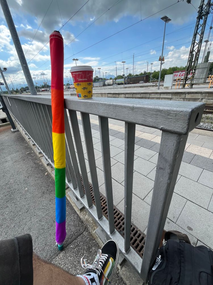 Eine große Regenbogenfahne lehnt an einem Geländer eines Bahnhofs. Auf dem Geländer steht noch ein Kaffeebecher. 