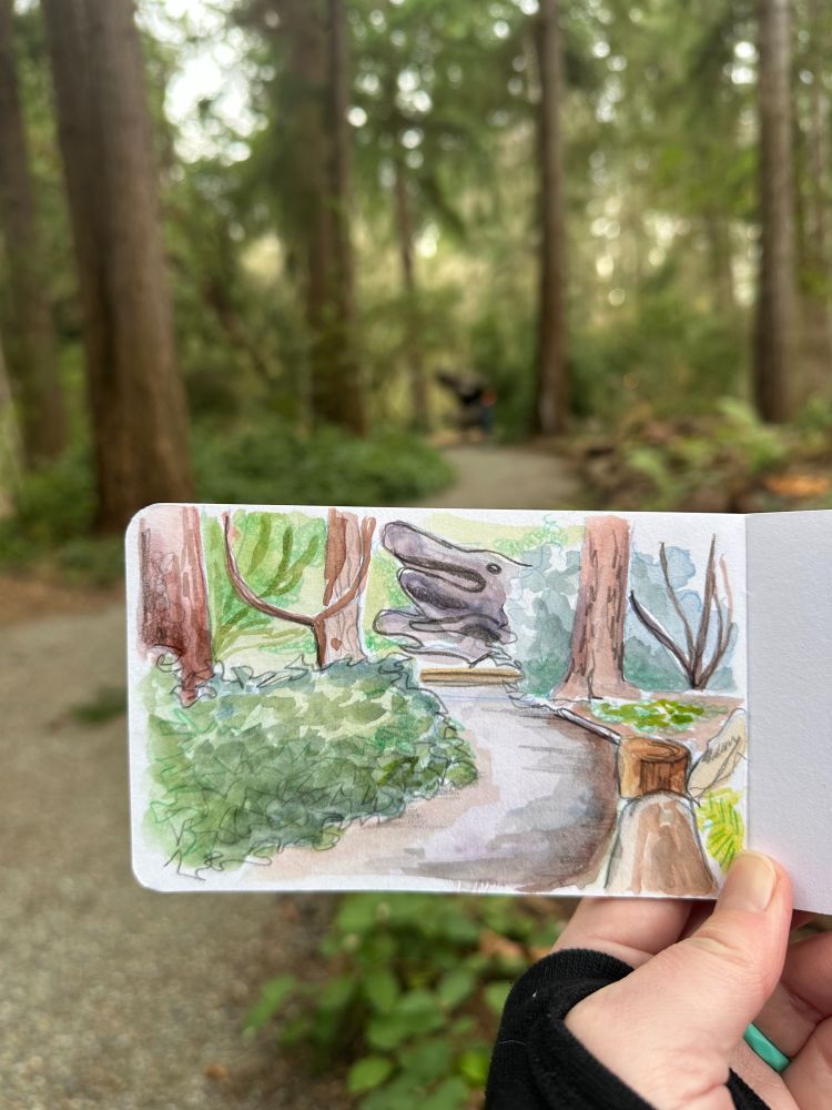 A sketchbook scene showing a garden path lined by logs on one side and greenery on the other. There's a large sculpture of tree roots in the background that looks sort of like a wolf snout. The sketchbook is held in front of the scene it is depicting.