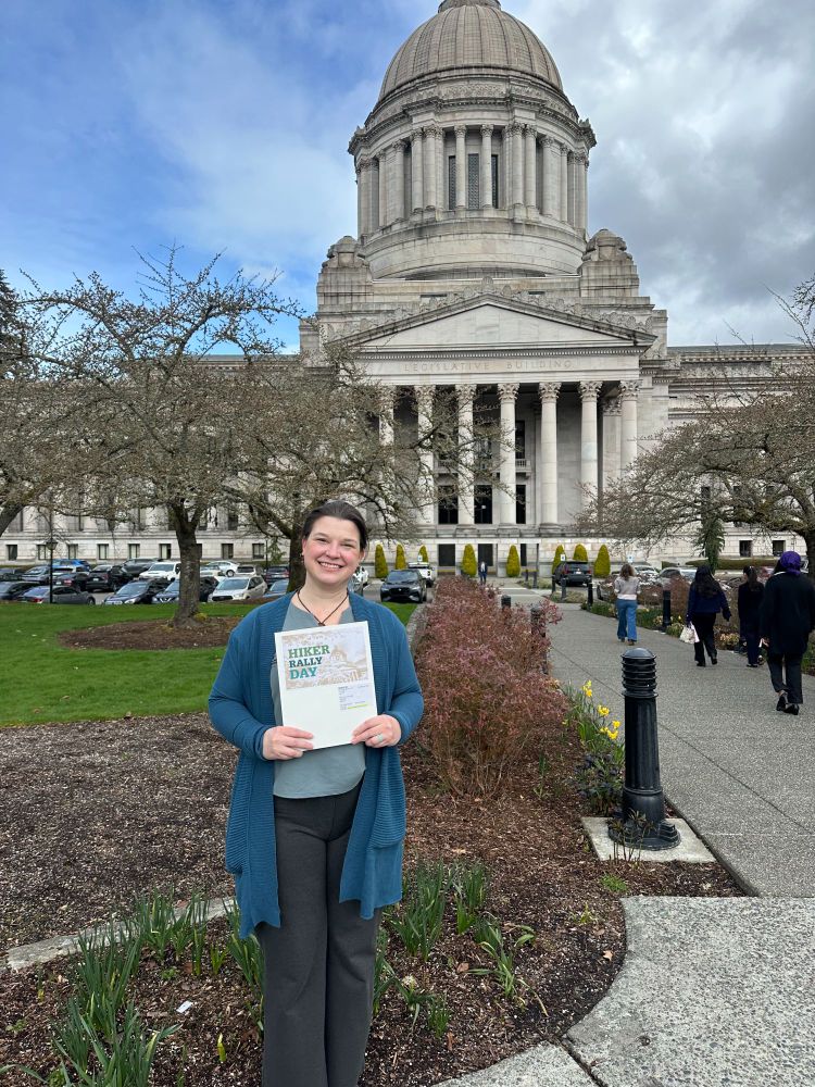 The artist holding up a folder labeled with the words "Hiker Rally Day" standing in front of the Washington state capitol building