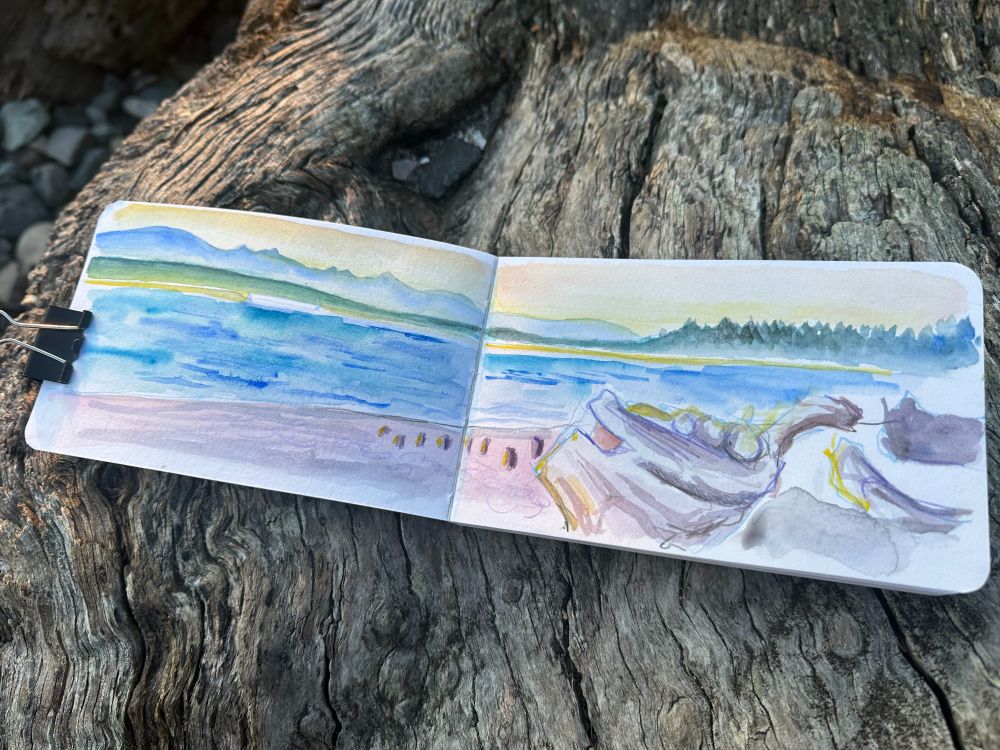 A small watercolor sketch of a Pacific Northwest beach at sunset. The sketchbook itself is sitting on weathered, gray driftwood.