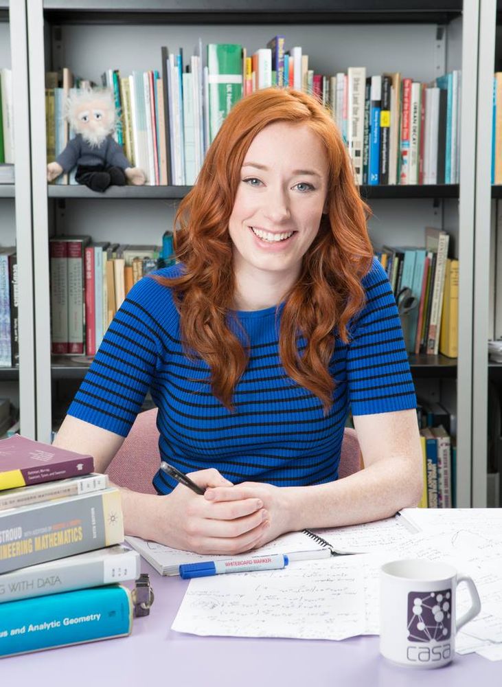 Hannah Fry, sitting at her desk. She is looking into the camera, a couple of mathematics books on the table.