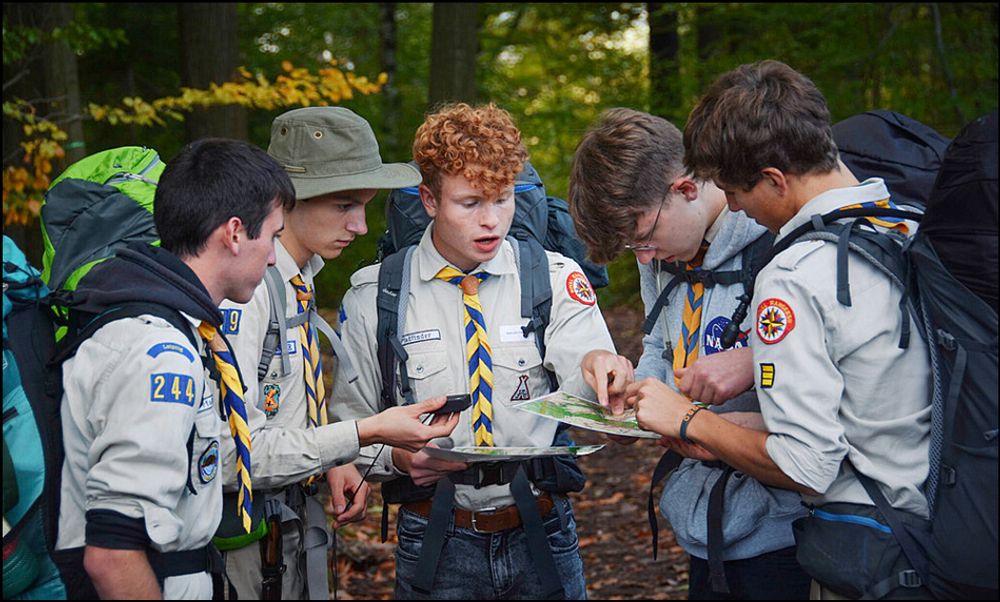 Zu sehen sind 5 männliche Jugendliche in Pfadfinderkleidung, die mittels Kompass und einer Landkarte, die Gegend erkunden oder das Ziel suchen. Sie stehen dicht beeinander und lösen gerade offensichtlich gemeinsam eine Schwierigkeit oder die Suche.