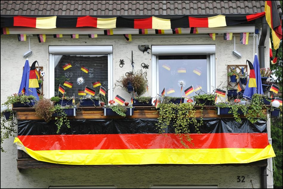 Zu sehen ist ein Balkon, der mit Deutschlandfahnen überladen ist und als Balkonbespannung dient eine riesengroße schwarz-rot-goldene Folie. Lauter Fahnen sind zu sehen, die auch an den Fenstern kleben. Unter dem Dach ist ein längerer Lappen in den Farben schwarz/rot/weiß zu sehen.